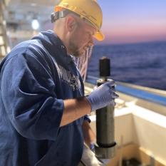 A white man in a blue shirt and a yellow hardhat walks from the left to right side of the images carrying a push core tube filled with ocean sediment