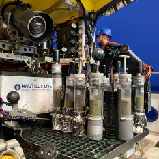 View of the front of ROV Hercules showing the yellow syntactic foam at the top of the image and a white sample box to the left of the picture. In the center is a black plastic milk crate on the flat extended porch of the vehicle. Inside and outside the crate are PVC sleeves holding vertical pushcore tubes filled about halfway with sediment. A dusky blue is visible behind the cores.