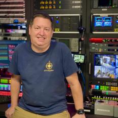 A man stands smiling in front of a wall of video equipment 