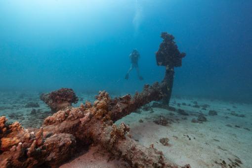 A diver floats midwater in tropical blue water behind a large ship's anchor laying in the sand